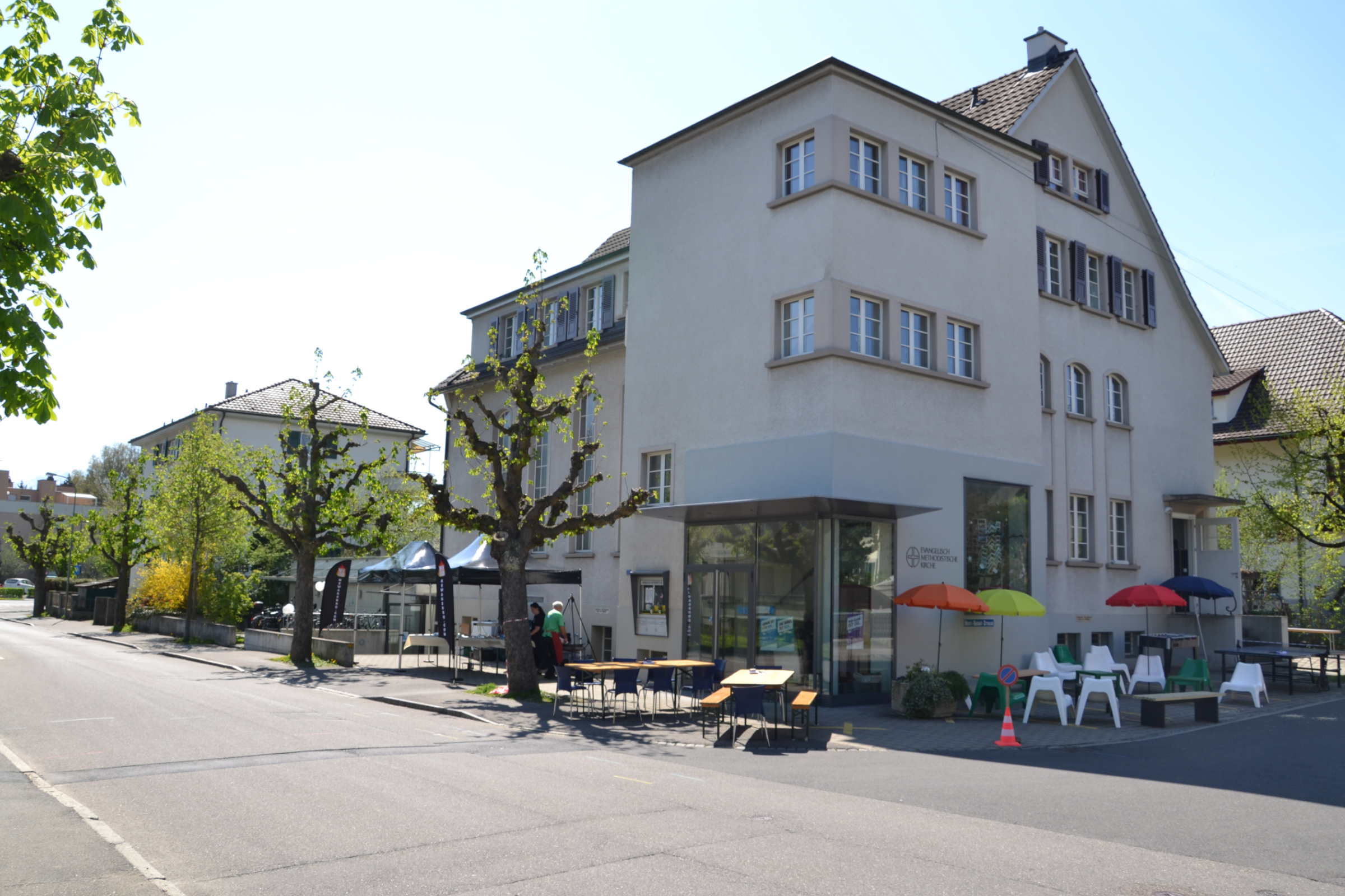 Vor der Kapelle an der Klosestrasse in Thun sind Partyzelte, Partytische, Loungesessel und Sonnenschirme aufgestellt. Es ist ein sonniger Tag, die ersten Blätter der Bäume beginnen zu treiben. Die Kapelle ist vierstöckig, ein eckiger Anbau im Vordergrund.